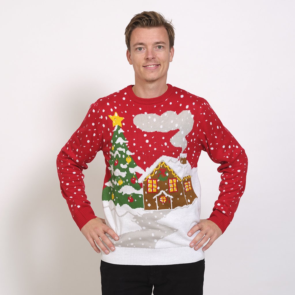 Un homme portant un pull de Noël rouge avec une maison enneigée et un sapin de Noël illuminé.